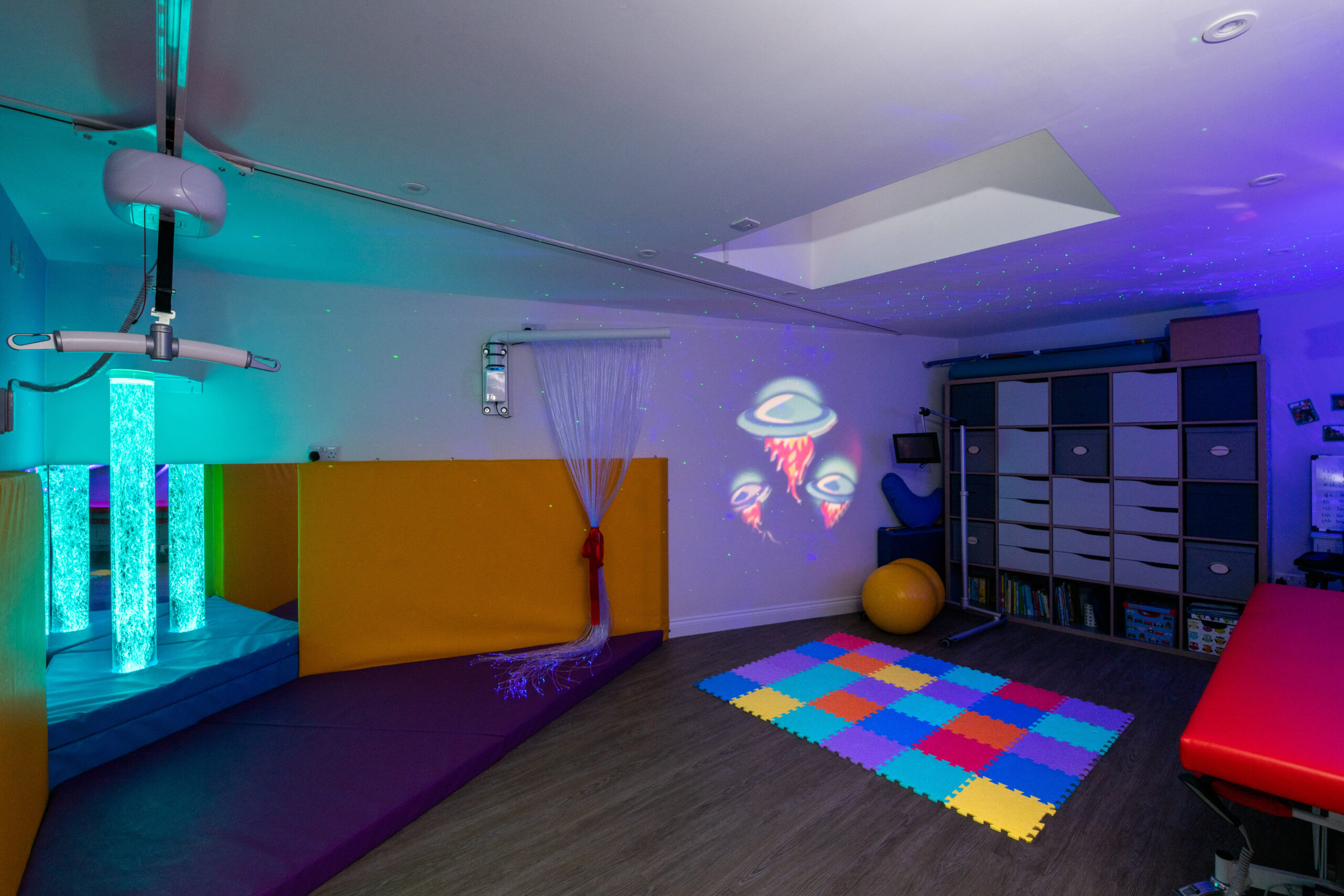 Accessible sensory room with ceiling hoist, bubble tube, fibre optic lighting, and soft play equipment designed for people with complex physical and sensory needs.